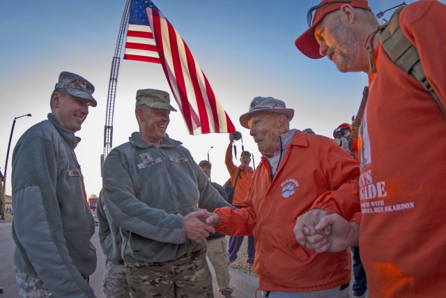 An American Flag hangs overhead as a senior marcher in an orange jacket shakes hands with one of two service members while others cheer in the background.